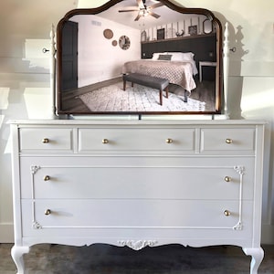 May include: A white dresser with gold knobs and ornate detailing. A large, arched mirror with a dark brown frame sits atop the dresser, reflecting a bedroom scene. The dresser has five drawers and curved legs.