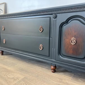 May include: A dark gray wooden sideboard with two drawers and two cabinet doors. The cabinet doors have a dark wood grain pattern. The drawers and doors have ornate bronze-colored hardware. The sideboard sits on short, turned wooden legs.