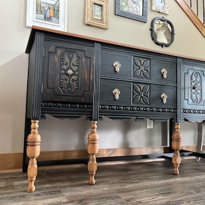May include: A black antique buffet with a light brown top and legs. The buffet has ornate carvings on the doors and drawers, with gold-colored hardware. The legs are turned and have a light brown finish.