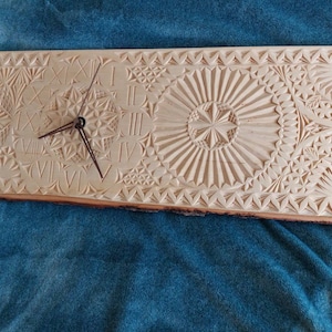 Może przedstawiać: Drewniany zegar z rzymskimi cyframi i misternymi rzeźbami. Tarcza zegara jest otoczona szczegółowym wzorem geometrycznych kształtów i wzorów.