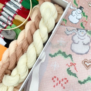 Könnte beinhalten: Eine weiße Box mit einem Kreuzstich-Set mit einem rosa und grünen Muster eines Schneemanns und einer Stechpalme. Das Set enthält Stickgarn in den Farben Creme, Beige und Braun.