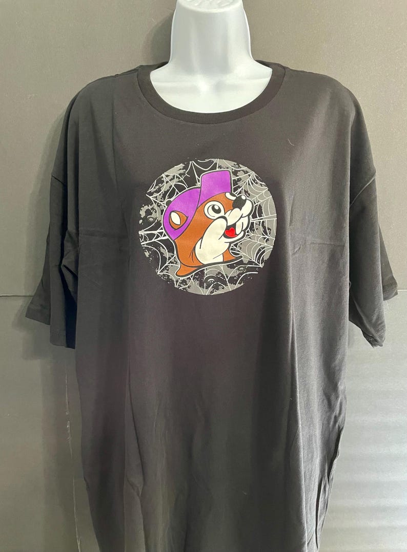 May include: Dark gray t-shirt featuring a cartoon beaver wearing a purple hat, set against a spiderweb design within a circle. The beaver has a friendly expression. The shirt is displayed on a white mannequin.