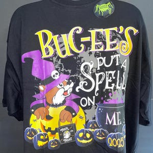 May include: Black Halloween t-shirt featuring a cartoon beaver in a witch hat holding a pumpkin. The shirt has the text "BUGEE'S PUT A SPELL ON ME" with a cauldron and jack-o'-lanterns. A glow-in-the-dark sticker is on the shoulder.