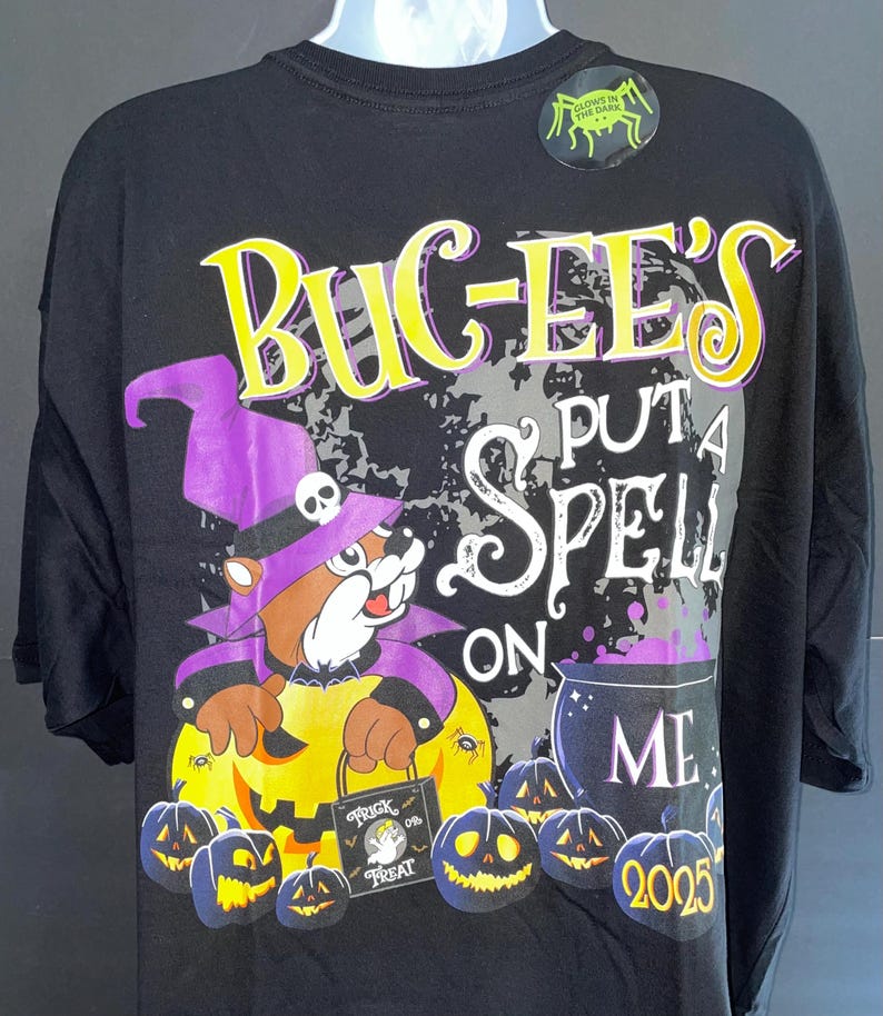 May include: Black Halloween t-shirt featuring a cartoon beaver in a witch hat holding a pumpkin. The shirt has the text "BUGEE'S PUT A SPELL ON ME" with a cauldron and jack-o'-lanterns. A glow-in-the-dark sticker is on the shoulder.
