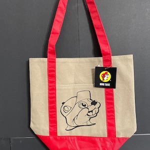 May include: A beige canvas tote bag with red handles and a red bottom. The bag features a black and white cartoon beaver graphic and a tag that reads "MINI TOTE".