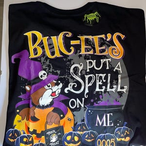 May include: Black t-shirt with a Halloween graphic. The design features a cartoon beaver in a witch hat, a cauldron, and jack-o'-lanterns. Text includes "BUC-EE'S", "PUT A SPELL ON ME", and "2025". A "Glows in the Dark" sticker is attached.