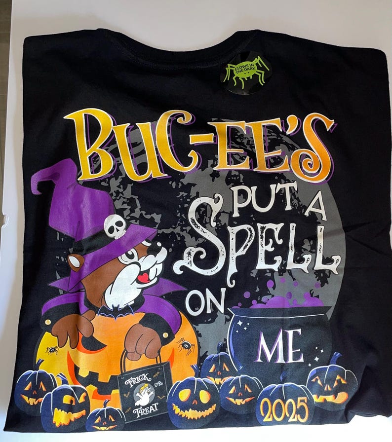 May include: Black t-shirt with a Halloween graphic. The design features a cartoon beaver in a witch hat, a cauldron, and jack-o'-lanterns. Text includes "BUC-EE'S", "PUT A SPELL ON ME", and "2025". A "Glows in the Dark" sticker is attached.