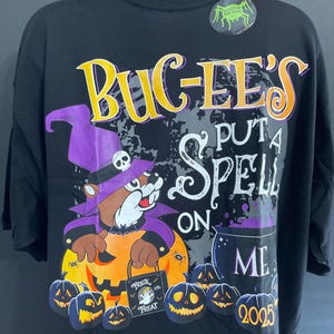 May include: Black Halloween-themed t-shirt featuring the text "Bug-ee's Put a Spell on Me" in yellow and white. The design includes a cartoon beaver in a witch hat holding a pumpkin, surrounded by jack-o'-lanterns and a cauldron. The year 2025 is also displayed.