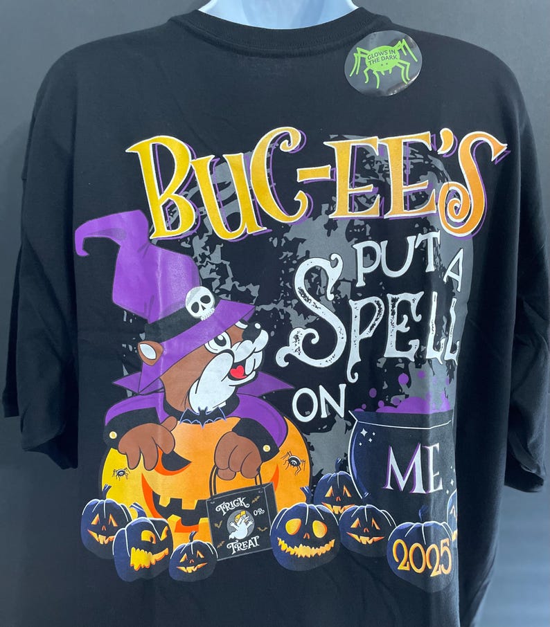 May include: Black Halloween-themed t-shirt featuring the text "Bug-ee's Put a Spell on Me" in yellow and white. The design includes a cartoon beaver in a witch hat holding a pumpkin, surrounded by jack-o'-lanterns and a cauldron. The year 2025 is also displayed.