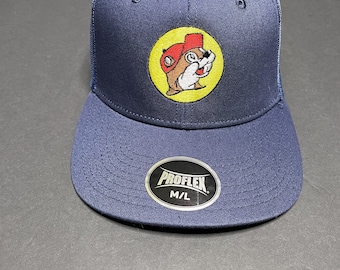 May include: Navy blue trucker hat with a mesh back and a curved brim. The front features an embroidered patch with a cartoon beaver wearing a red hat. The hat has a ProFlex M/L label.