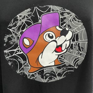 May include: Black sweatshirt featuring a cartoon beaver wearing a purple hat. The beaver is brown, white, and has a red heart-shaped tongue. The design is set against a gray spiderweb background with small ghost-like figures.