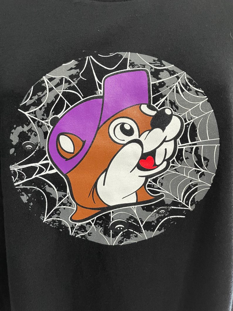 May include: Black sweatshirt featuring a cartoon beaver wearing a purple hat. The beaver is brown, white, and has a red heart-shaped tongue. The design is set against a gray spiderweb background with small ghost-like figures.