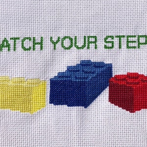 Könnte beinhalten: Kreuzstich-Stickerei mit dem Text "WATCH YOUR STEP" in grünem Garn und einem grünen Herzen. Unter dem Text befinden sich drei farbige Lego-ähnliche Blöcke: gelb, blau und rot.