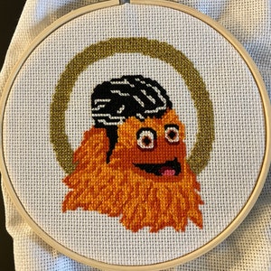 May include: A cross-stitch embroidery of a cartoon character with orange fur, a black and white striped hat, and a big smile. The character is surrounded by a gold circle on a white background.
