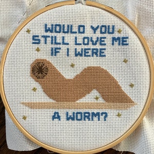May include: A white embroidery hoop with a brown worm embroidered on it. The worm has a brown eye and is surrounded by gold stars. The text "Would you still love me if I were a worm?" is embroidered in blue thread.