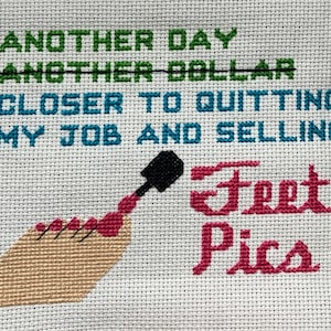 Op de afbeelding: Borduurwerk met kruissteek met de tekst "Another day, another dollar, closer to quitting my job and selling feet pics". Het ontwerp bevat een hand met rode nagellak en een flesje nagellak.