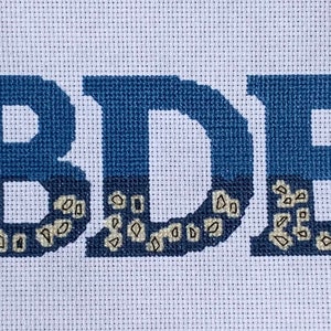 Puede incluir: Un bordado de punto de cruz azul y blanco con las letras "BDE" en una fuente de bloque.