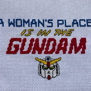 May include: A white cross-stitch embroidery with the text "A woman's place is in the Gundam" in blue, yellow, and red thread. A yellow and white Gundam robot head is embroidered below the text.