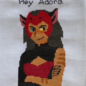 Pode incluir: Bordado de ponto cruz de um personagem de desenho animado com cabelo vermelho, uma faixa de cabeça vermelha e uma roupa preta. O personagem tem um olho azul e um olho amarelo. O texto "Hey Adora" está bordado acima do personagem.