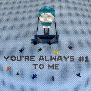 May include: A blue and white cross-stitch design featuring a hot air balloon with a blue and white bus underneath. The text "You're always #1 to me" is stitched in gray below the balloon.