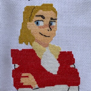 Op de afbeelding: Borduurwerk met kruissteek van een cartoonfiguur met blond haar, die een rood jasje en een wit shirt draagt. De tekst "Hey Catra" staat boven het figuur.