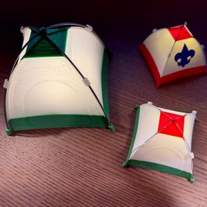 Pode incluir: Três objetos em miniatura em forma de tenda. O maior é branco com acabamentos e detalhes verdes e hastes de suporte pretas. Os outros dois são menores, com detalhes vermelhos e verdes. Um tem um quadrado vermelho no topo e o outro uma flor-de-lis azul.