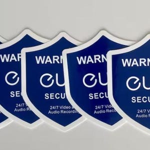 May include: A set of blue and white security warning stickers. Each shield-shaped sticker displays the text "WARNING!! eufy SECURITY" and "24/7 Video and Audio Recording". The stickers are designed to indicate video surveillance.
