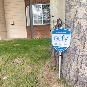 Puede incluir: Un letrero de seguridad azul y blanco con el texto "GUARDED BY eufy SECURITY 24/7 Audio and Video Surveillance" está sujeto a un tronco de árbol. El letrero está frente a un edificio con revestimiento marrón y una ventana.