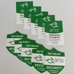 May include: A set of green and white stickers with the Arlo logo and the text "Monitoring in Progress" and "Security Video Monitoring in Progress".