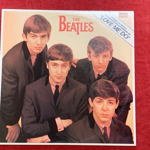 May include: Album cover featuring The Beatles, with the band members in suits. The band's name is in red, with the title "LOVE ME DO" on a white banner. The image has a vintage aesthetic.