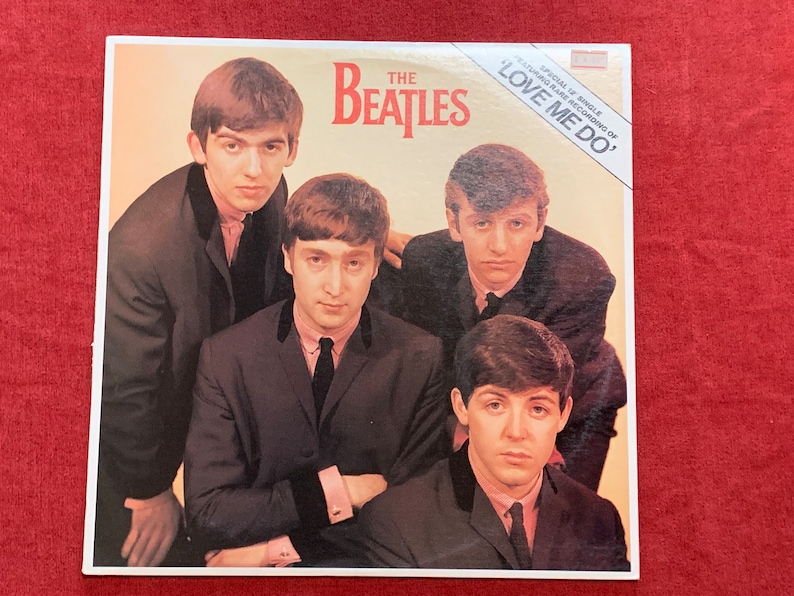 May include: Album cover featuring The Beatles, with the band members in suits. The band's name is in red, with the title "LOVE ME DO" on a white banner. The image has a vintage aesthetic.