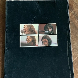 May include: Vintage black book cover with four framed images of The Beatles. The images show the band members with microphones. The cover has a worn appearance, with visible wear and tear. A collectible item.