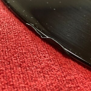 May include: Close-up of a black, glossy object with a damaged edge, resting on a red, textured surface. The object's surface reflects light, showing subtle scratches and imperfections. The damaged edge reveals a white interior.