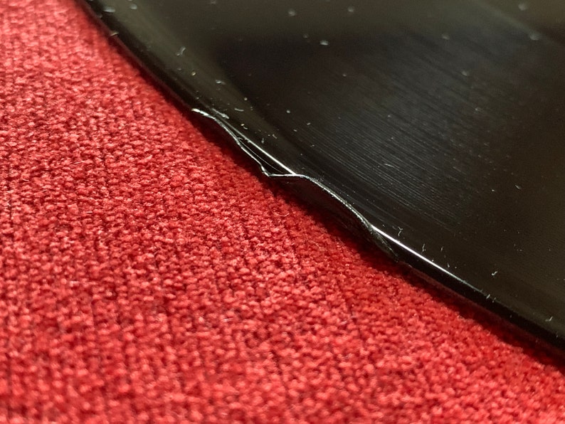 May include: Close-up of a black, glossy object with a damaged edge, resting on a red, textured surface. The object's surface reflects light, showing subtle scratches and imperfections. The damaged edge reveals a white interior.