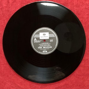 May include: A black vinyl record with a white and gray label featuring the text "THE BEATLES" and song titles "LOVE ME DO" and "P.S. I LOVE YOU." The record is set against a red background.
