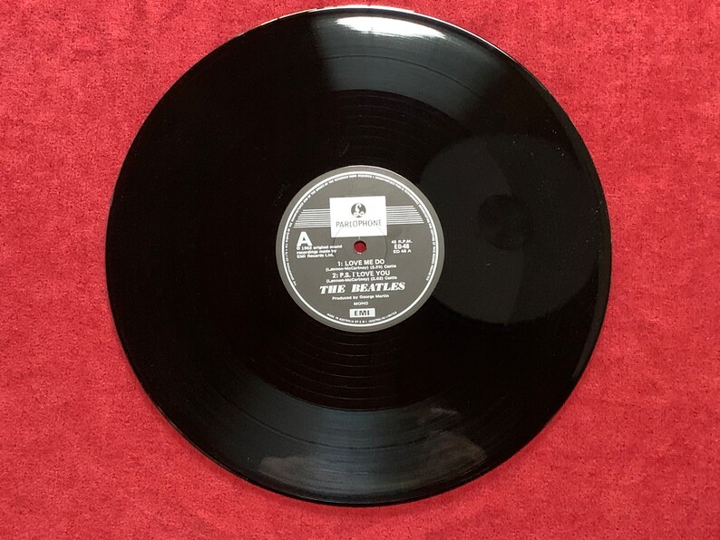 May include: A black vinyl record with a white and gray label featuring the text "THE BEATLES" and song titles "LOVE ME DO" and "P.S. I LOVE YOU." The record is set against a red background.
