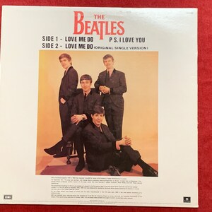 May include: Album cover for The Beatles' single, featuring the band members in black suits. The text reads "SIDE 1 - LOVE ME DO, SIDE 2 - LOVE ME DO, P.S. I LOVE YOU." The background is a light orange color.