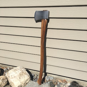 May include: A wooden-handled axe with a black metal head. The axe is leaning against a light brown wall with a wooden texture.