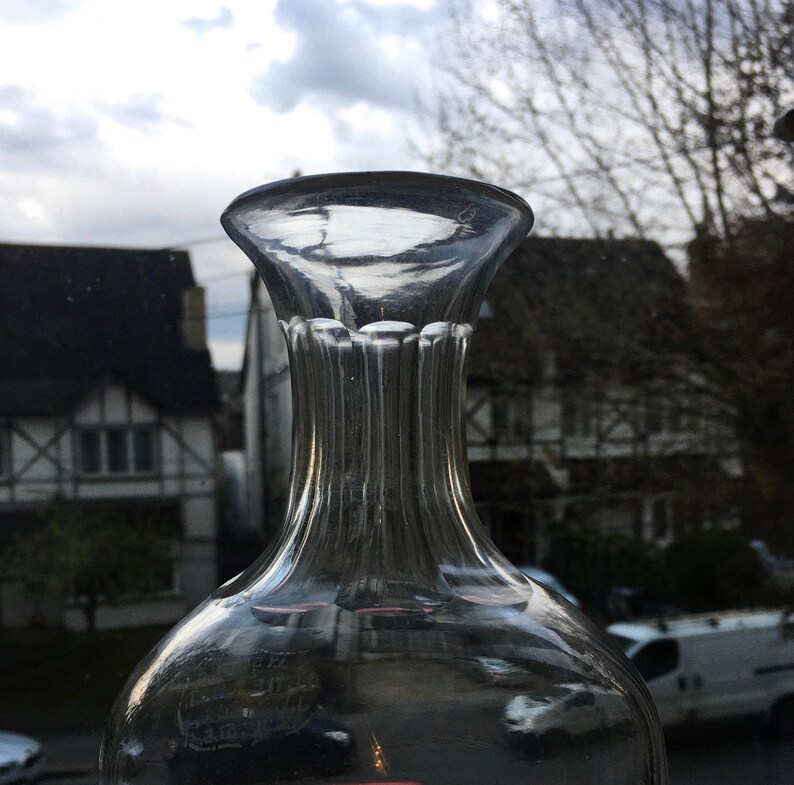 May include: A clear glass decanter with a wide mouth and a textured, ribbed neck. The decanter is empty and reflects the surrounding environment.