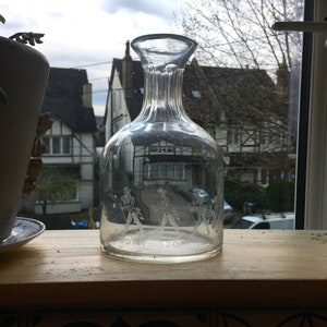 May include: A clear glass decanter with a textured surface and a wide mouth. The decanter has an etched design of three figures in formal attire. The text "JOHN 320" is etched on the bottom of the decanter.