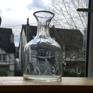 May include: A clear glass decanter with a textured surface and etched figures. The decanter is sitting on a wooden surface. The text on the decanter reads "Little John, Much the Miller's Son, Able & Strong".