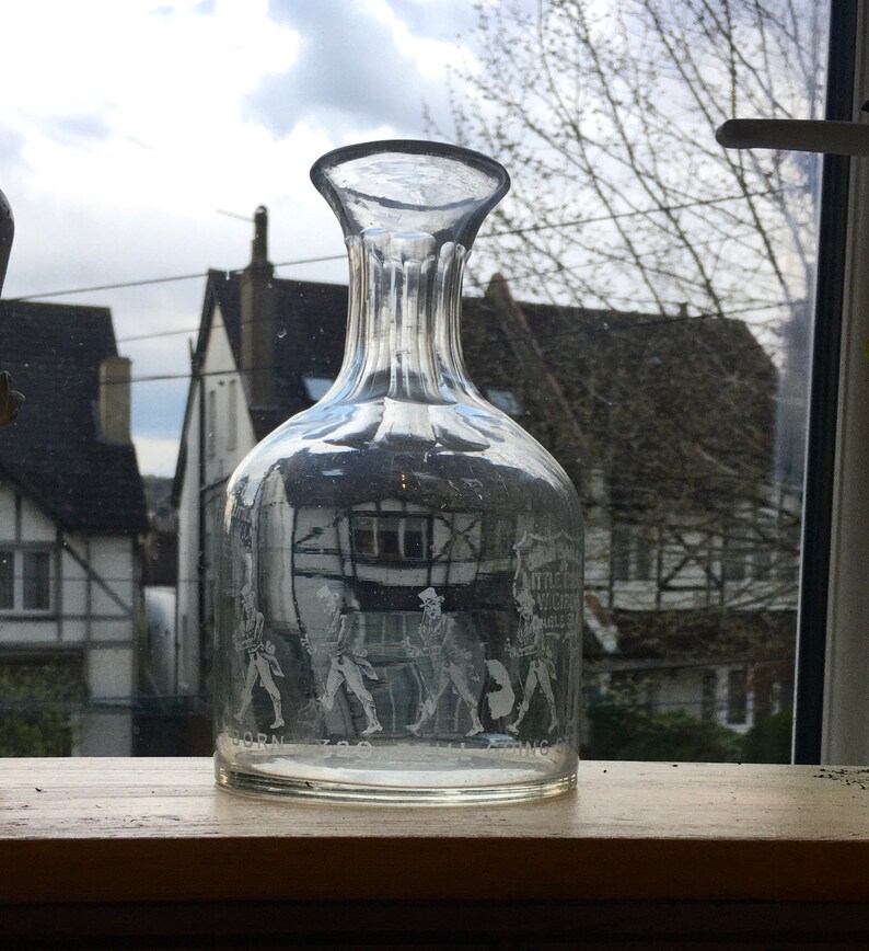 May include: A clear glass decanter with a textured surface and etched figures. The decanter is sitting on a wooden surface. The text on the decanter reads "Little John, Much the Miller's Son, Able & Strong".