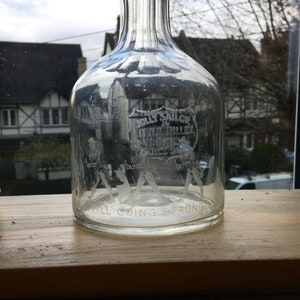 May include: A clear glass decanter with a textured neck and a label that reads "Jolly Sailor Little Bullet Whiskey Charles St. Still Going Strong".