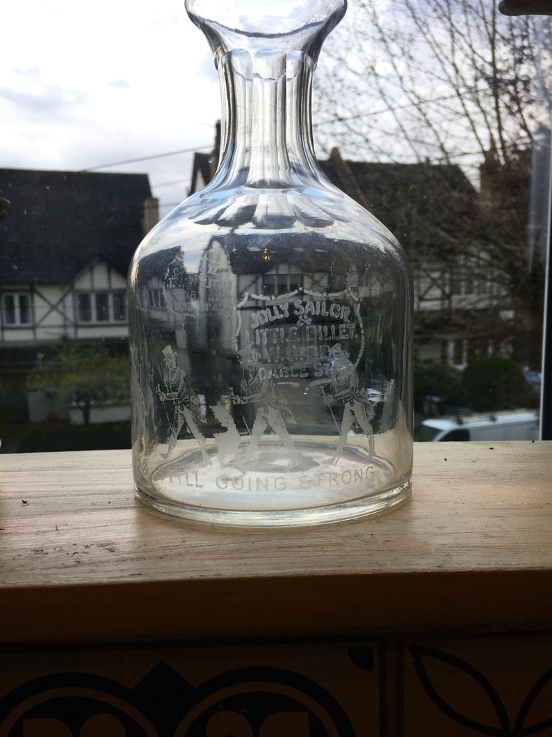 May include: A clear glass decanter with a textured neck and a label that reads "Jolly Sailor Little Bullet Whiskey Charles St. Still Going Strong".