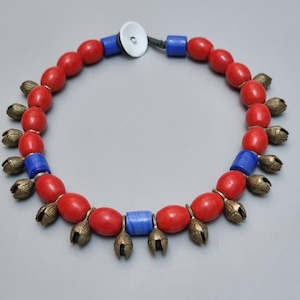 May include: A necklace featuring red oval beads, blue cylindrical beads, and small brass bells. The necklace has a white circular clasp and a dark cord. The beads are evenly spaced.