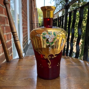 Puede incluir: Un jarrón de vidrio rojo con detalles dorados y flores pintadas. El jarrón tiene una base ancha y un cuello estrecho.