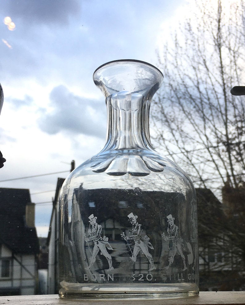 May include: A clear glass decanter with a textured surface and a wide mouth. The decanter has three figures etched into the glass, each wearing a hat and holding a walking stick. The text "BORN. 320. STILL GO" is etched at the bottom of the decanter.