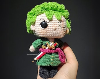 Op de afbeelding: Een groene gehaakte figuur met een zwart ooglapje en een rood-witte sjerp. De figuur draagt een groen-gouden gehaakt shirt en een zwarte broek.