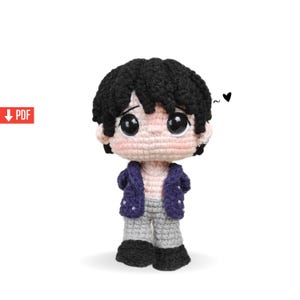Puede incluir: Muñeca de ganchillo hecha a mano con cabello negro, ojos negros grandes y rostro rosa claro. La muñeca viste una chaqueta azul, pantalones grises y zapatos negros. Un pequeño corazón negro está dibujado encima de la cabeza de la muñeca.