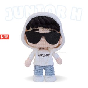 May include: A crocheted doll wearing a white hoodie, sunglasses, and blue jeans. The doll has black hair and the text "$AD BOY" on the front of the hoodie. The doll is standing on a white surface.